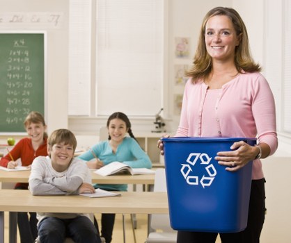 In a classroom setting, a woman with shoulder-length light brown hair, wearing a light pink top, stands near the front holding a blue rubbish bin with a white recycling symbol printed on it. Behind her, three children are seated at light-colored desks, with two girls and one boy, all smiling and facing forward. The background features a white wall with a green chalkboard on the left side displaying handwritten mathematical equations or numbers, and windows allowing natural light to illuminate the room. The scene appears to be part of an educational environment focused on promoting cleanliness and recycling, which aligns with rubbish removal services like those offered by Office Clearance Southwark operating in the Southwark area. The overall atmosphere is bright and orderly, with a focus on environmental awareness and waste management education.