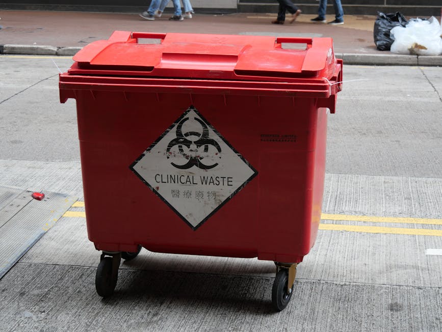 A person wearing a dark blue jacket is placing used AA batteries into a green battery recycling container situated on a table or counter. The individual holds the batteries between their thumb and ind