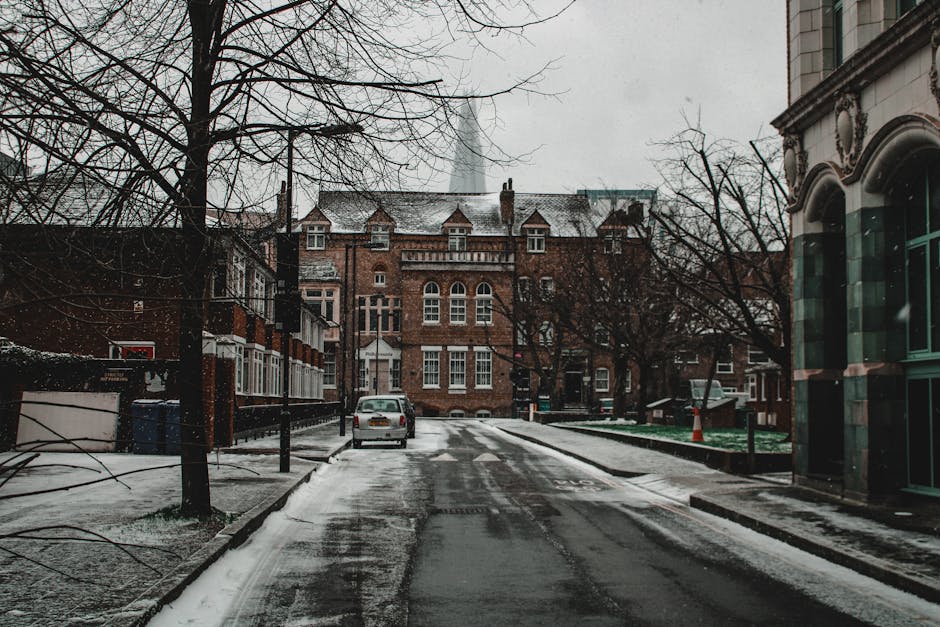The image depicts an urban street scene featuring a weathered building facade with a small, white-framed window partially covered by a roller shutter, situated above a row of three large waste bins po