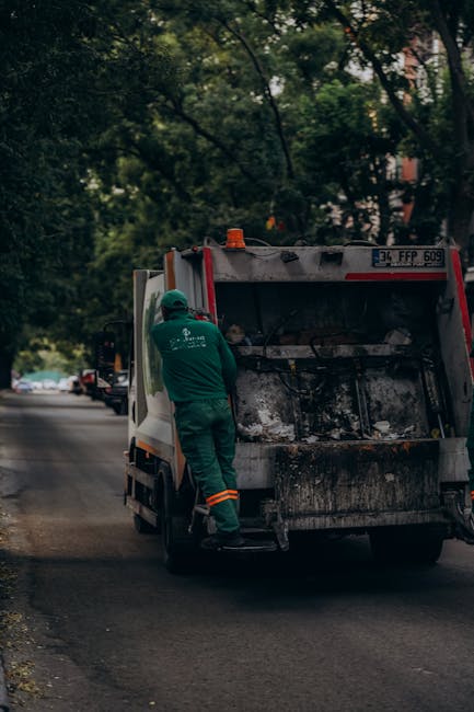 A narrow urban alleyway is filled with a large, wheeled trash bin in the foreground, which appears to be overloaded and covered with a worn, gray fabric sack. Behind it, a collection of discarded card
