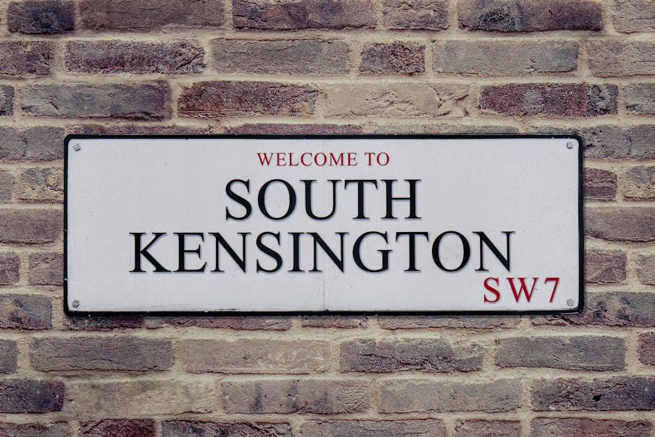 A rectangular white metal street sign mounted on a reddish-brown brick wall. The sign features the text 'WELCOME TO SOUTH KENSINGTON' in black capital letters, with 'WELCOME TO' in smaller red font ab